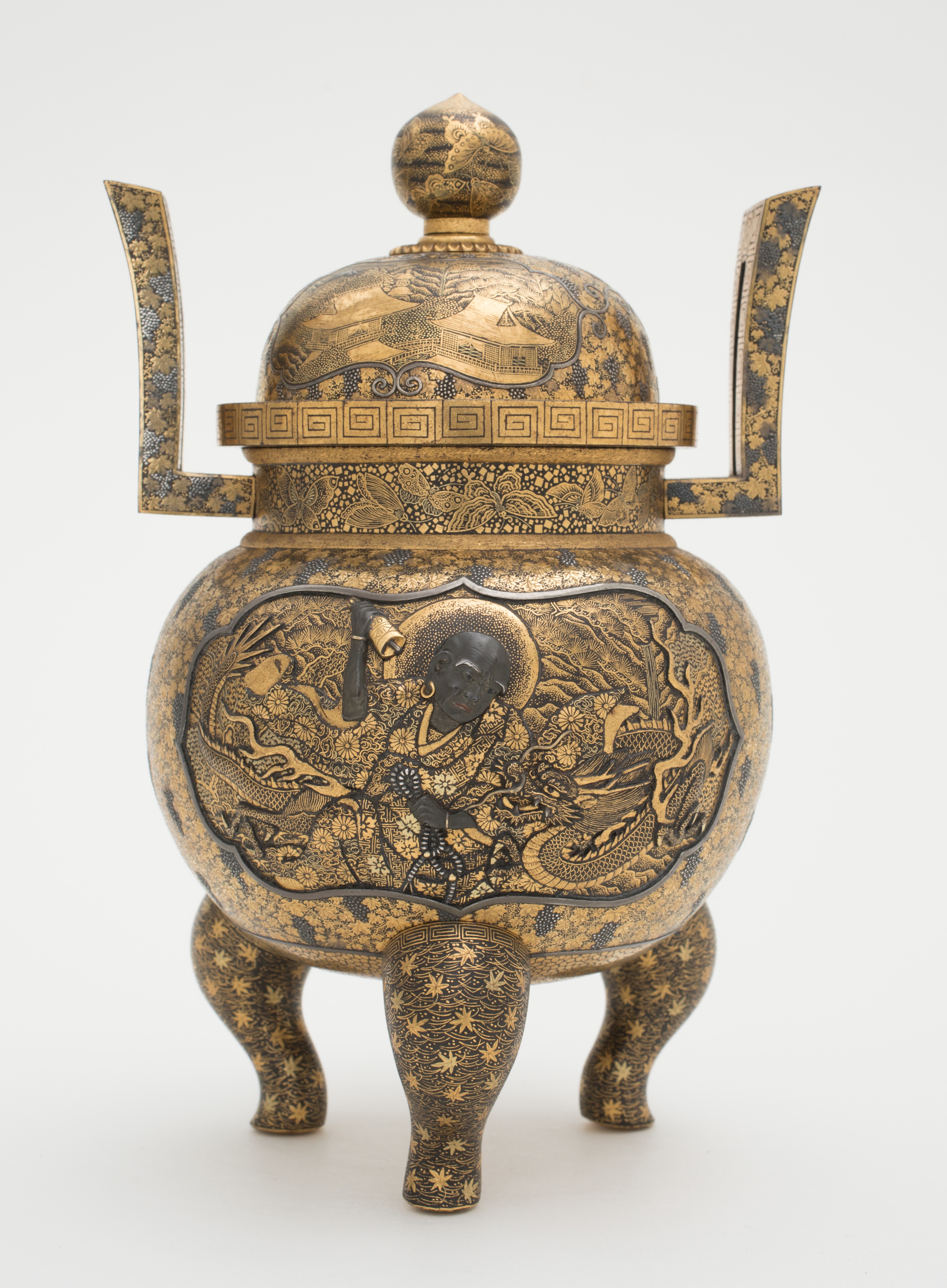 Three legged koro with lid and upright handles on either side. On the belly of the incense burner is a dragon and a budda who is holding a bell in one hand and a string of beads in his other